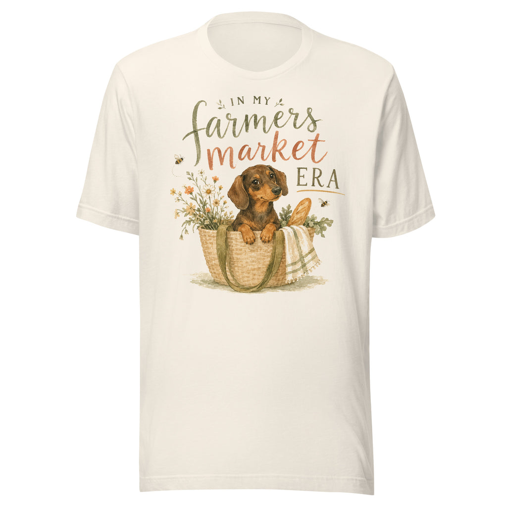 Cream unisex t-shirt reading In My Farmers Market Era with a watercolor illustration of a dachshund puppy in a market tote basket with flowers and bread
